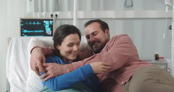 Happy Young Man Visiting and Cheering Woman Lying in Bed at Hospital Ward