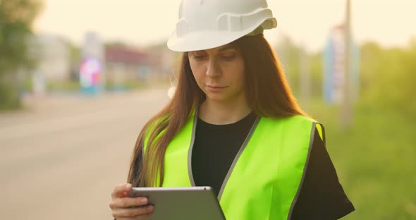 Female Inspector in Light Green Vest and Protective Helmet with Tablet in Her Hands Monitors the