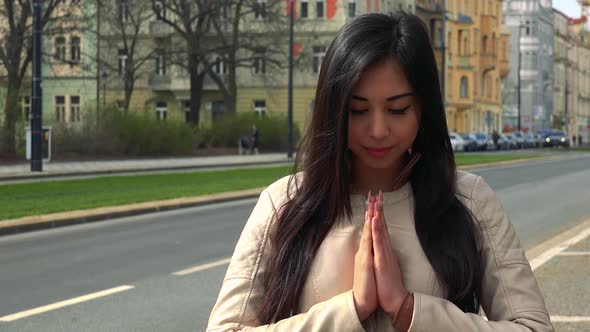 A Young Asian Woman Prays with Hands Clasped Together in a Street in an Urban Area