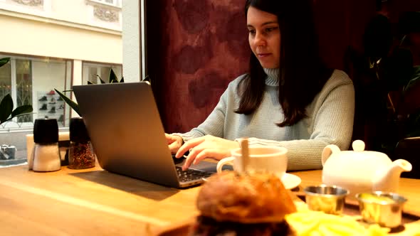 Woman Working Remotely on Laptop in Cafe Eating Burger Drinking Tea