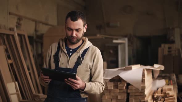 A Young Man with a Beard Walks Around the Carpentry Shop with a Tablet in His Hands