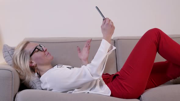 Young Blonde Woman with Glasses Is Lying on Couch and Calling By Video To Friends Using Smartphone