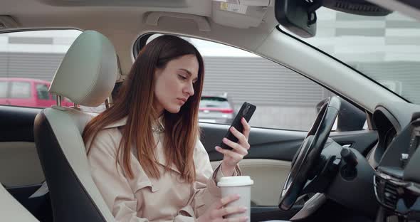 Elegant Woman Sitting in Car and Surfing Feed News on a Smartphone and Drinking Coffee