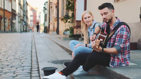 Young Couple of Street Singers Sitting on Sidewalk Playing Guitar and Singing