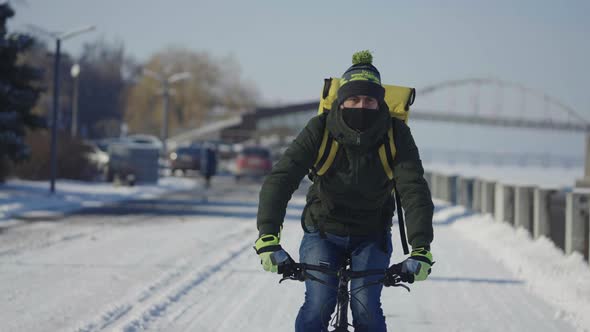Portrait of Cyclist with Backpack and Protective Mask Rides Bicycle on Snow Covered Street with Food
