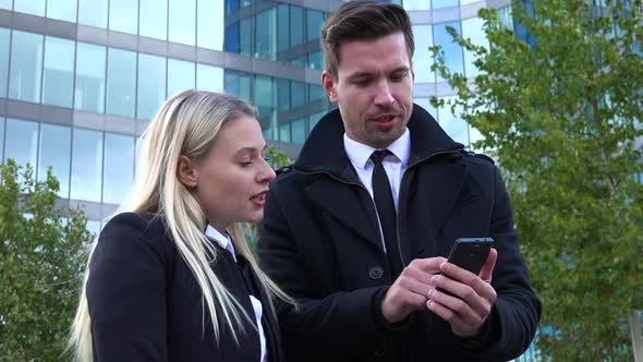 A Businessman and a Businesswoman Work on a Smartphone and Talk - an Office Building in Background