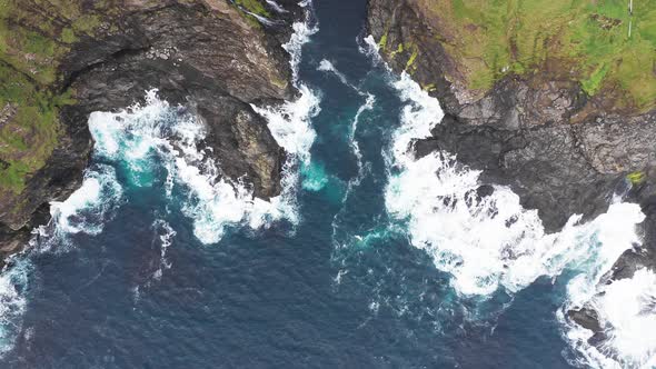 Aerial View of Waves Break on Rocks of Faroe Islands Cliffs in a Blue Ocean