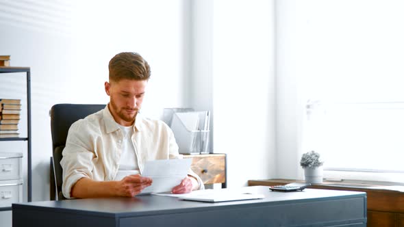 Skilled bearded accountant in yellow shirt looks through reports on sheets of papers