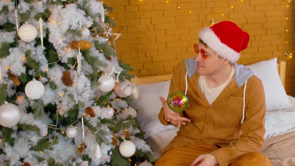Portrait of Man in Santa Hat and Bright Glasses with Disco Bauble Sitting on Bed Near Christmas Tree