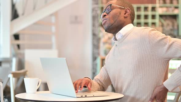 Hardworking African Man with Laptop Having Back Pain in Cafe