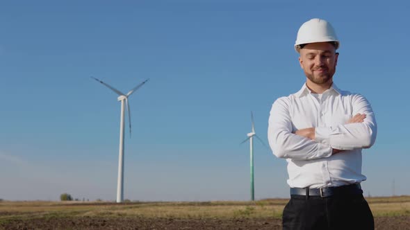 Air Power Engineer Stands in Business Style Clothes and White Helmet on a Background of Windmill