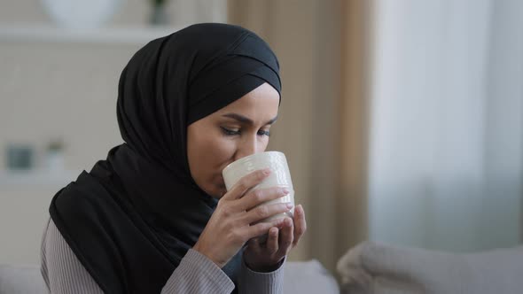 Carefree Joyful Muslim Girl in Hijab Drinking Tea Relaxing in Room Alone Look Thoughtfully Into