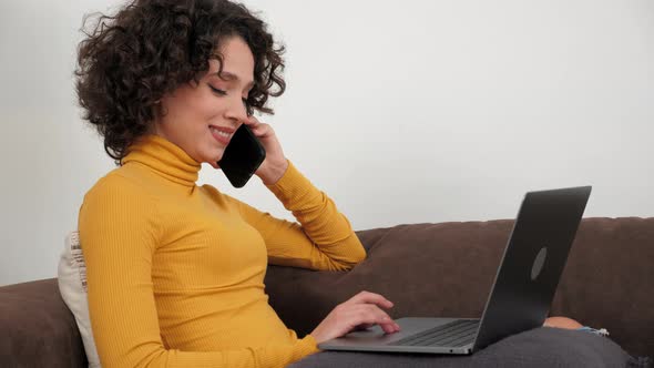 Smiling Hispanic Businesswoman Using Laptop Listens on Mobile Phone at Home