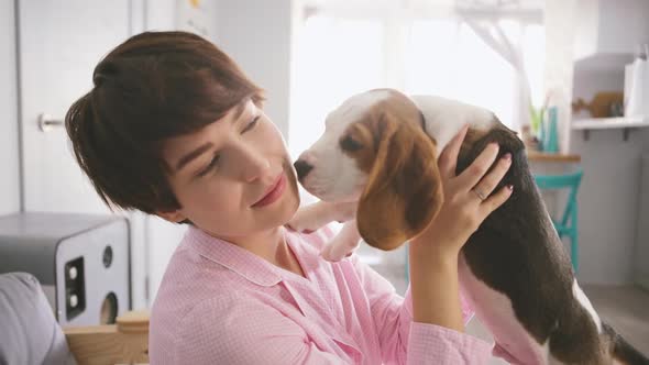 Young Caucasian Female in Pink Pyjamas Holding Beagle Puppy in Arms and Kissing Him in Slowmotion