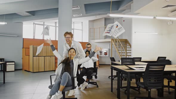 Young Multiracial Diverse Colleagues are Throwing Up Documents or Papers While Racing on Chairs in