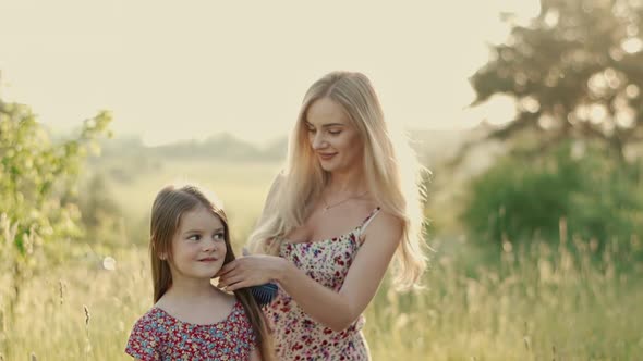 Smiling Blonde Mom in a Summer Dress Combs Her Daughter's Hair