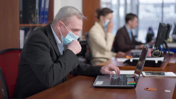 Side View of Ill Senior Caucasian Man in Coronavirus Face Mask Coughing with Blurred Coworkers