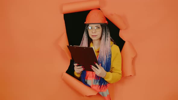 Female construction worker holding clipboard and sticking out of hole of orange background.