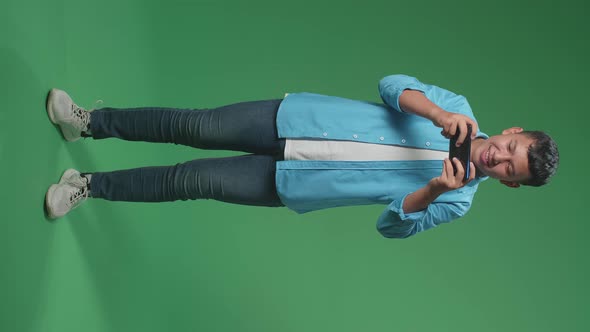 Full Body Of Excited Young Asian Boy Playing Video Game With Mobile Phone In Green Screen Studio