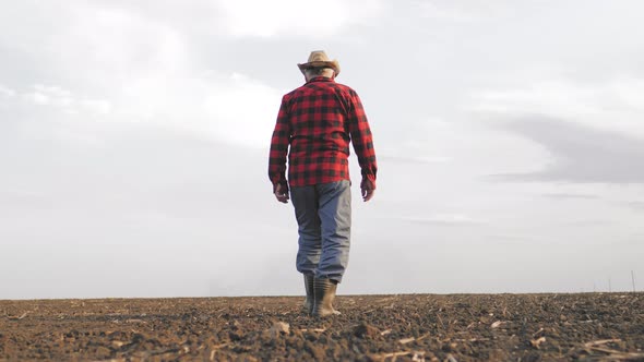 Senior Male Farmer Walks Through a Field with Plowed Land in His Rubber Boots