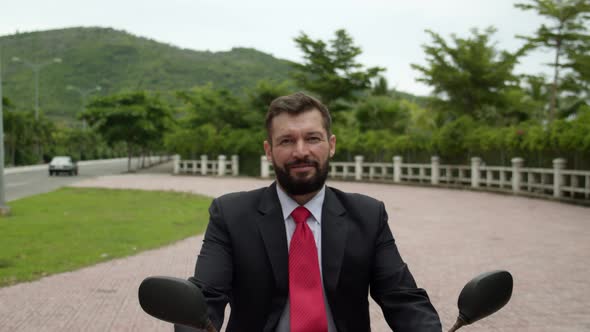 Portrait of a Senior Bearded Smiling Businessman Driving Up on a Motorcycle Along the Street