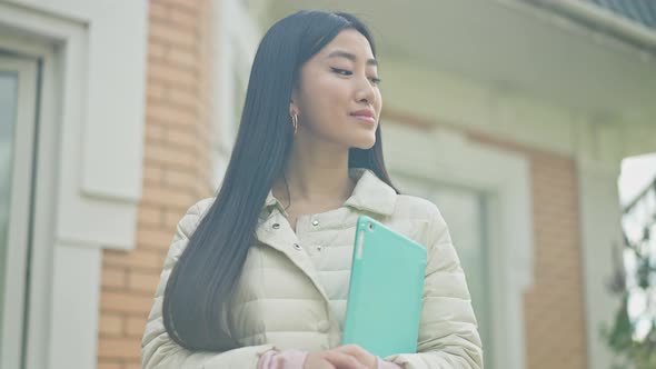 Middle Shot of Smiling Asian Millennial Standing on House Porch with Tablet in Sunlight