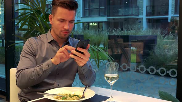 A Young, Handsome Man Sits at a Table in a Restaurant and Works on a Smartphone