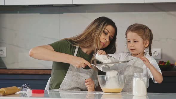 Mother and Girl Sift Flour with Sieve at Table in Kitchen