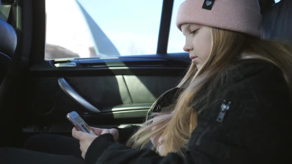 Beautiful Female Child with Long Blonde Hair Uses Her Smart Phone During Riding on Modern SUV