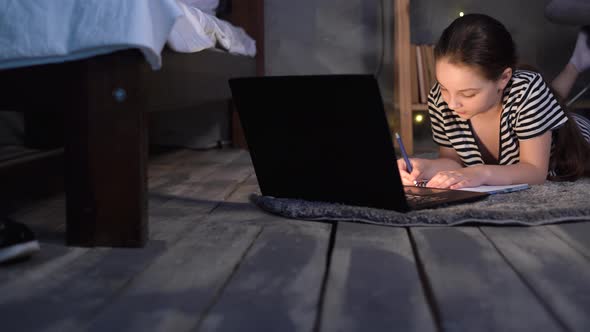 Teenage Girl Doing Homework Using Laptop Lying on Bedroom Floor in Evening Female Student Writing alt
