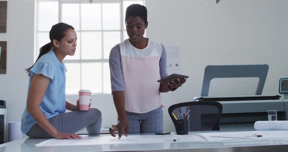 Two diverse businesswoman holding tablet and coffee, working together in office