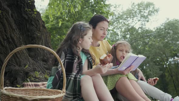 Mature Grandmother Sit in the Park Near Tree on the Blanket Reading a Book with Two Granddaughters