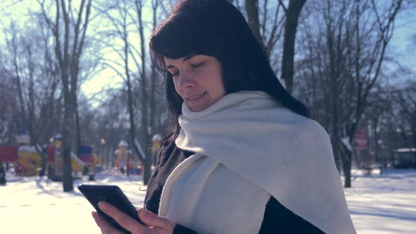 Happy Smiling Beautiful Girl Using Smart Phone. Sunny Day In Snow Covered City Park. Slow Motion