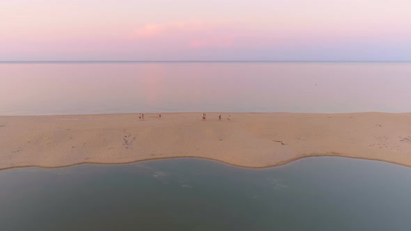 Group of People Walking on Sand Stripe at Sea Coast Against Flat Sea Horizon at Sunset with Pink