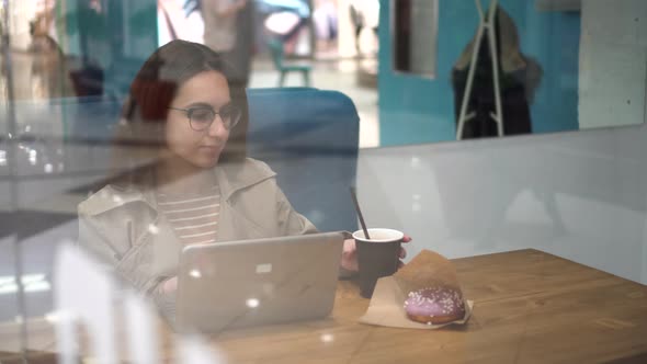 Young Woman Sitting Behind the Glass in a Cafe with a Laptop