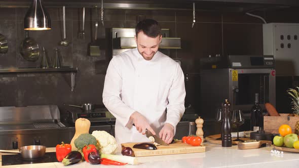 Young Chef Slices Eggplant on a Wooden Board in the Interior of Modern Restaurant's Kitchen