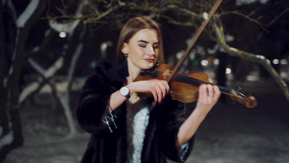 A talented girl in warm clothes is performing composition on the violin in a winter park