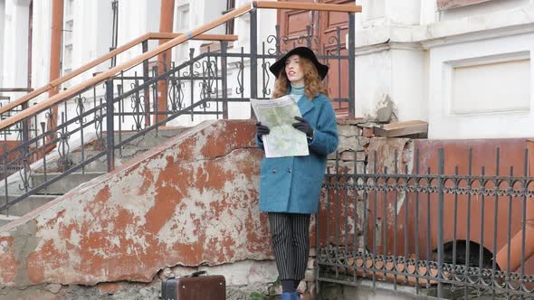 Young Redhaired Woman in a Blue Coat and Black Hat with a Suitcase on the Background of the Old City