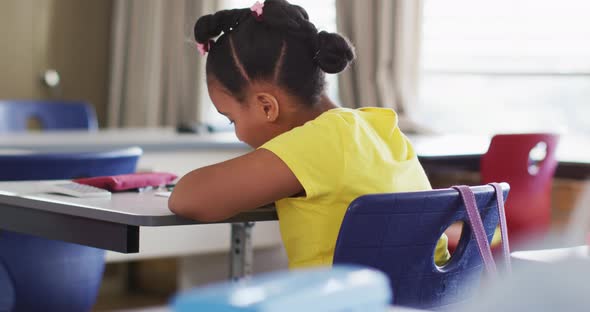 Portrait of happy mixed race schoolgirl sitting at classroom, making notes, looking at camera