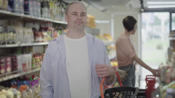 Positive Male Buyer Posing with Shopping Basket in Grocery As Blurred Woman Choosing Goods at the