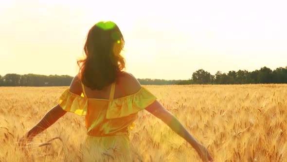 Young Woman in Yellow Dress Standing on a Wheat Field with Sunrise on the Background