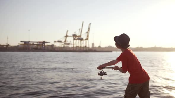 Young Caucasian Boy in Hat and Red Tshirt Standing Near Sea with Port on Background Throwing Bait in