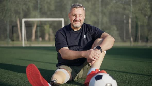 Portrait of Confident Smiling Handsome Sportsman Amputee Sitting on Summer Stadium Looking at Camera