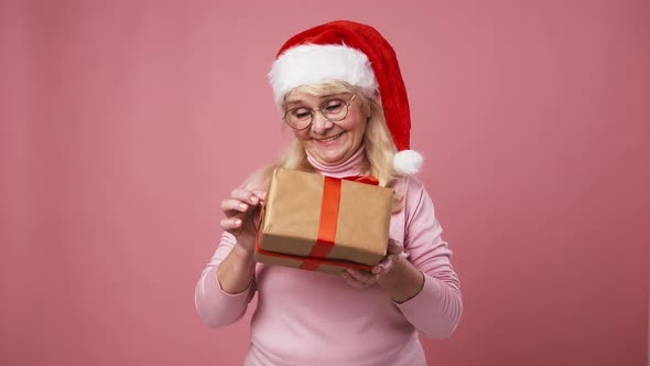 Happy Senior Woman in Santa Hat Getting Gift and Shaking Box Guessing What's Inside Pink Background