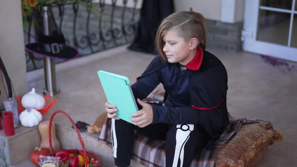 Cute Boy in Vampire Skeleton Costume Watching Movie Online on Tablet Smiling in Slow Motion Sitting