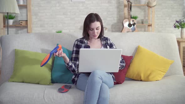 Young Woman Sitting on the Couch Uses a Laptop and Holds Orthopedic Insoles in Her Hand