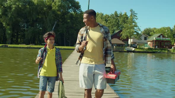 Happy African Father and Teenage Boy with Rods Walking on Wooden Pier on Lake