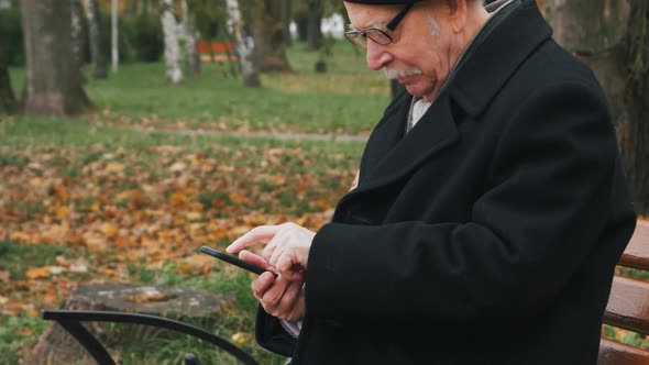 Elderly senior sitting on a bench and using smartphone or cell phone in park. Tapping, scrolling, wa