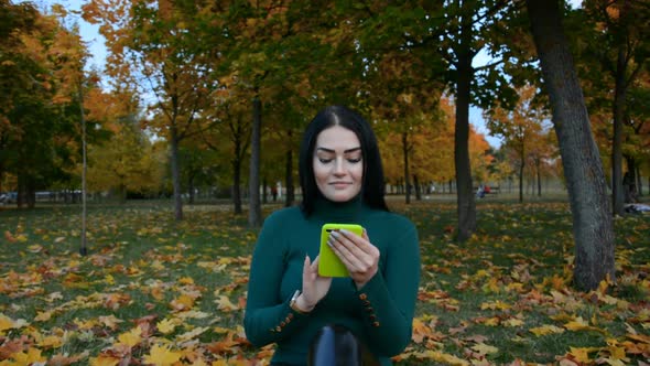 A Beautiful Girl is Sitting in an Autumn Park on the Grass and Texting with Friends on Social