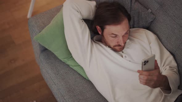 Confident long-haired man looking at phone while lying on the sofa